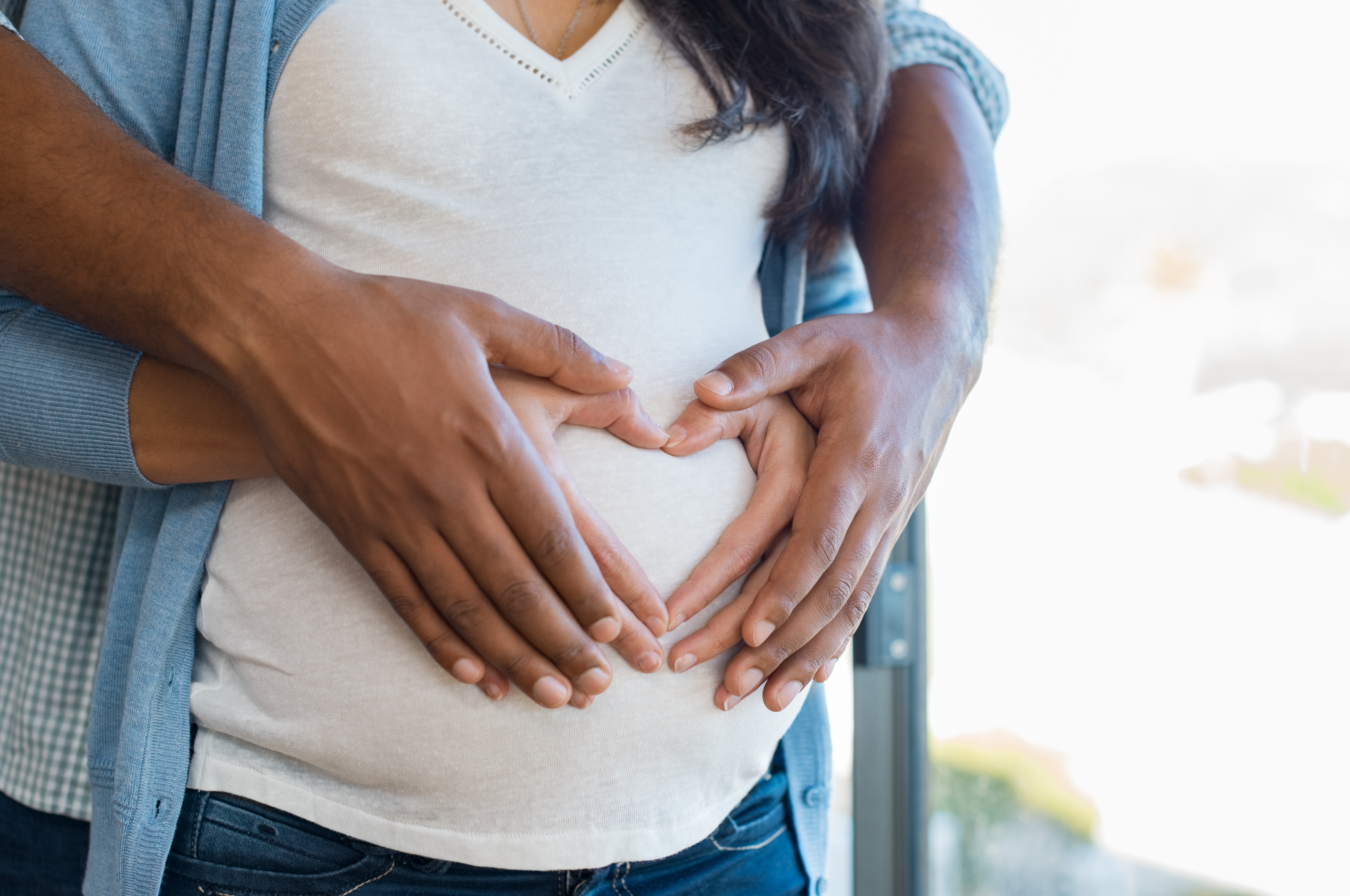 Pregnant woman and her african husband holding hand in heart shape on baby bump. Close up of multiethnic couple making heart shape on the tummy. Loving future couple expecting a baby.